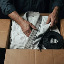 Hands folding a gi and a black belt into a cardboard box, ready to post in