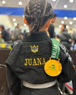 JUANA and a crown embroidered in gold on a child's black gi, worn with a competition medal
